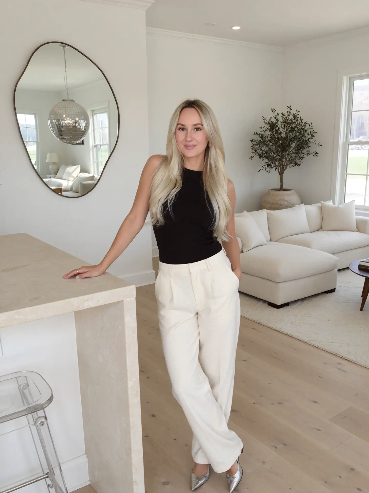 AI Twin in a black top and cream trousers leaning at a marble kitchen island